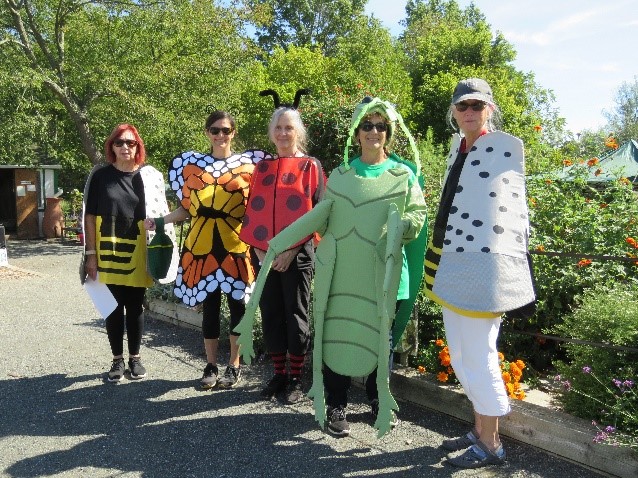 Four women in insect costumes pose in front of a garden