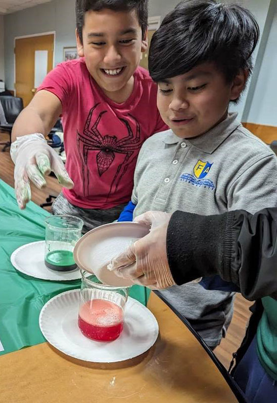 Two smiling young boys look on as a gloved hand pours salt into test tubes of liquid one red and one green. 