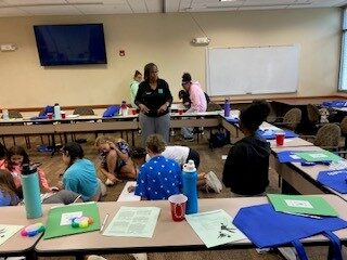 Woman stands in middle of room teaching to a group of young people