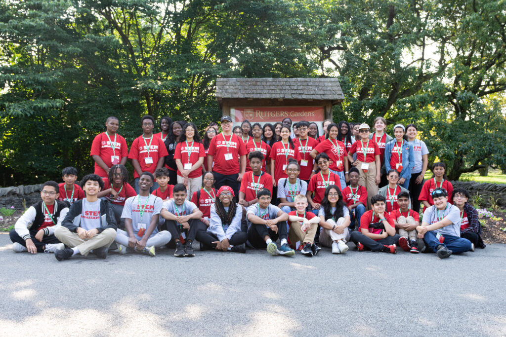 Photo shows the cohort of the STEM Ambassadors for the 2023 - 2024 year. Teens wearing either red or gray shirts in front of a Rutgers Garden sign