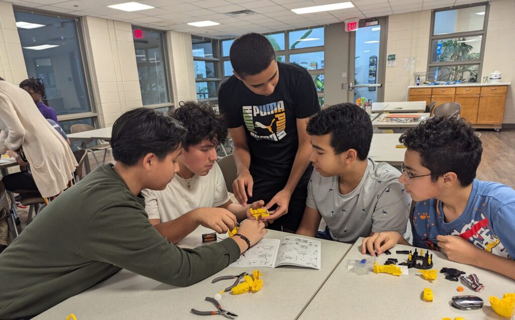 4 sitting youth look on while a youth that is standing holds a yellow object. There are tools and other yellow objects on a table in front of the youth. 