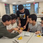 4 sitting youth look on while a youth that is standing holds a yellow object. There are tools and other yellow objects on a table in front of the youth.