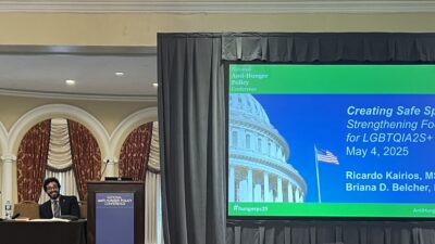 Picture of a large screen with the words National Anti-Hunger Policy Conference to the left are a man and a woman behind a long table.