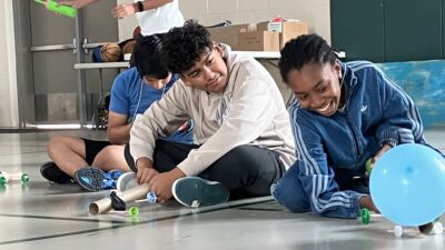 Three youth's watch as a balloon powered car is released in a gym