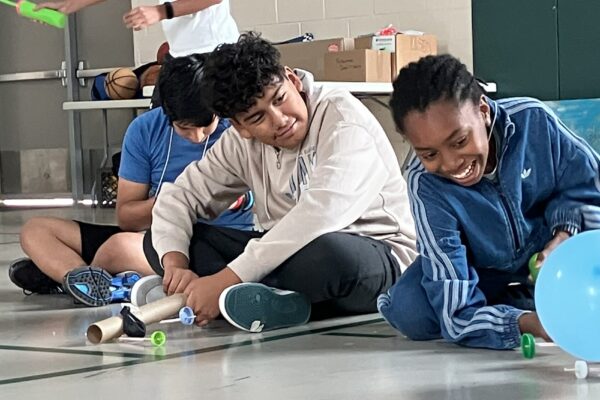 Three youth's watch as a balloon powered car is released in a gym