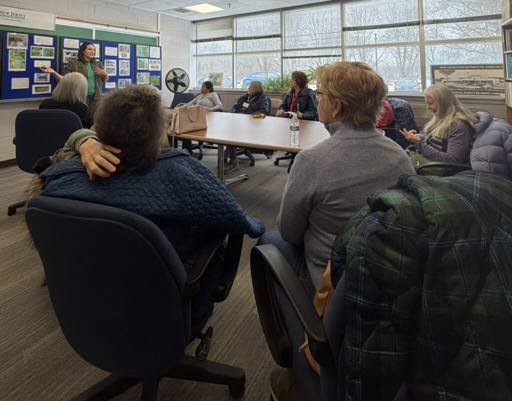 A group of women sit around a table watching as a woman in the front of the room talks while pointing at a display.
