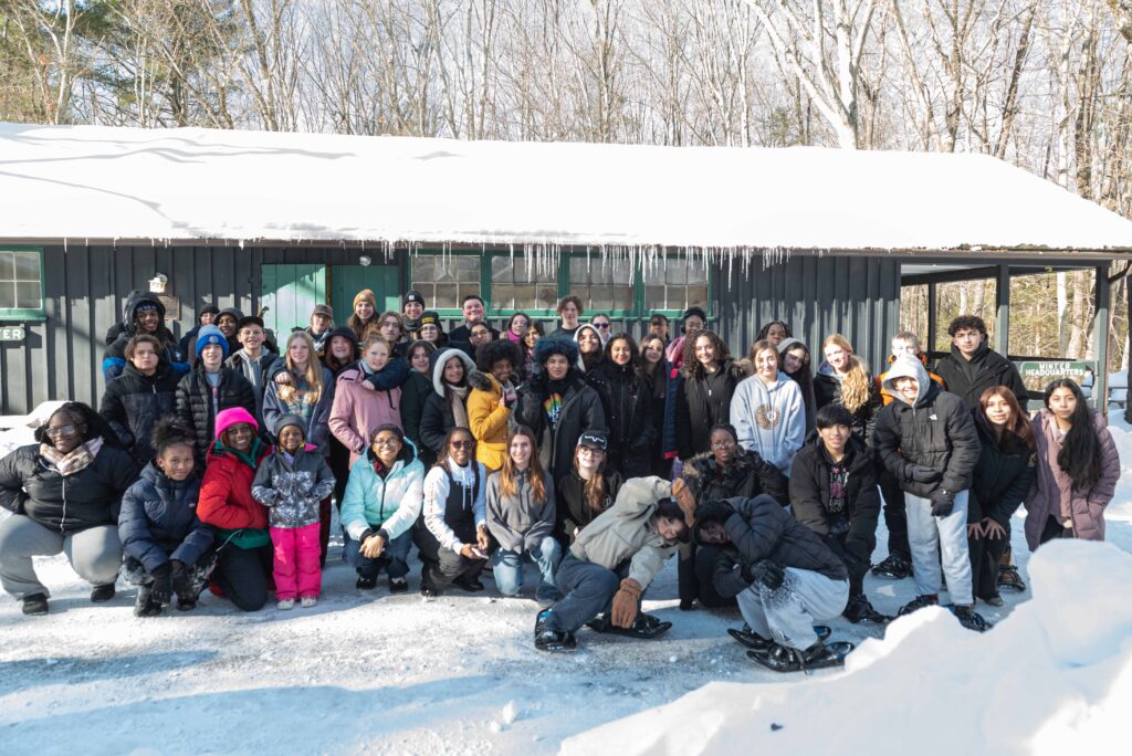 a large group of teens clad in winter gear are gathered in front of an icicle covered house . There is snow on the gound and on the roof.