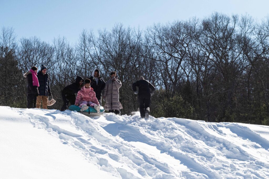 Three teens stand at the top of a snow covered hill. One teen is on a sled and is being pushed down the hill by another teen. A person with their back towards the camera walks up the hill. Two adults stand nearby watching the teens.