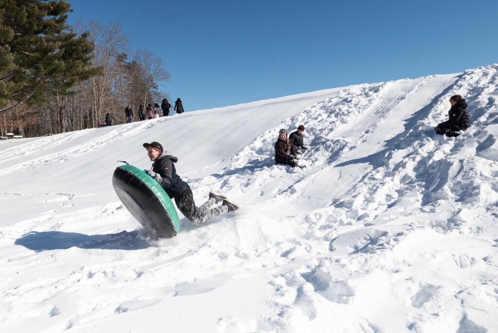 A laughing teen is on a snow tube sledding down a snow covered hill while three other teens look on. In the distance at the top of the snow covered hill there is a group standing and looking on.