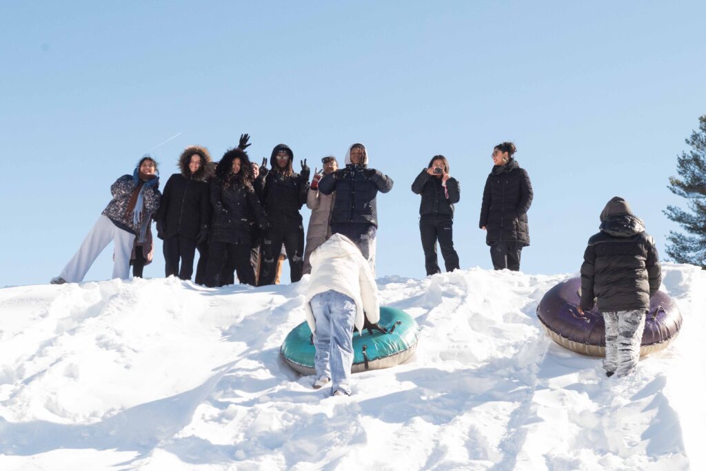 Ten teenagers stand on top of a snow covered hill smiling for the camera as two young people with their back towards the camera push sleds up the hill
