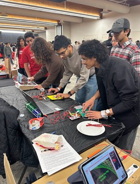 a long table with a series of people working with computers and screens in the background, teens with STEM Ambassador shirts look on