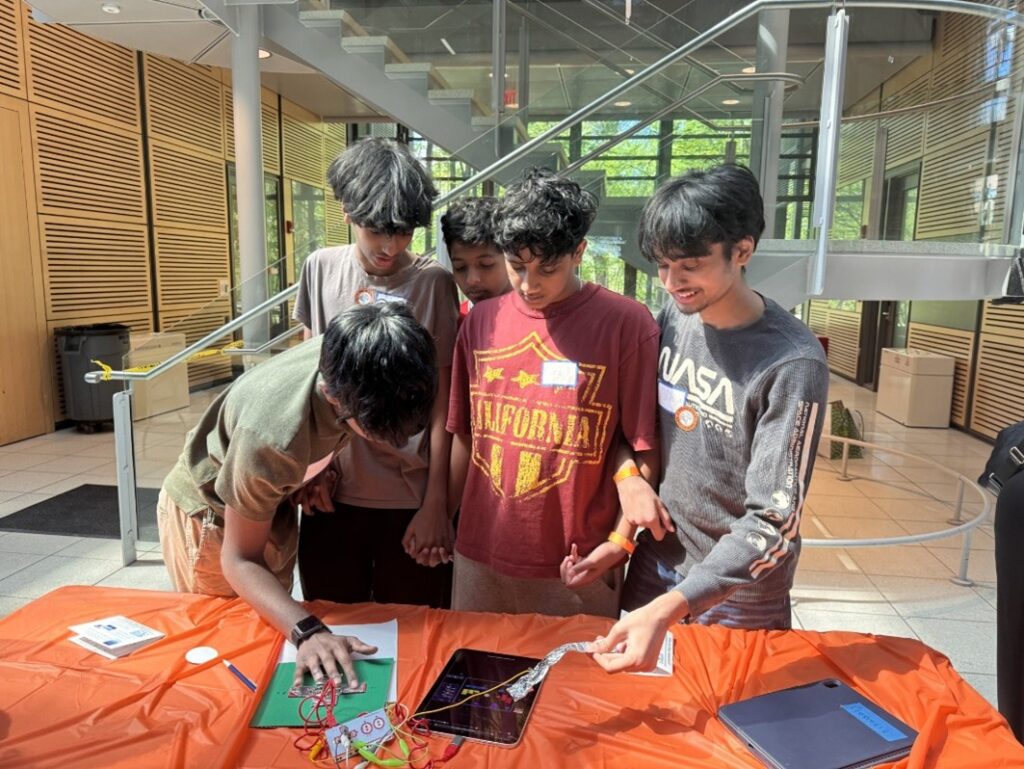 5 young teens gather around an iPad, connecting wires and concentrating on the screen