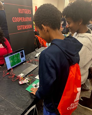 Two young boys gather around a computer with a soccer field on the screen, playing a game with colorful pads connected to the computer